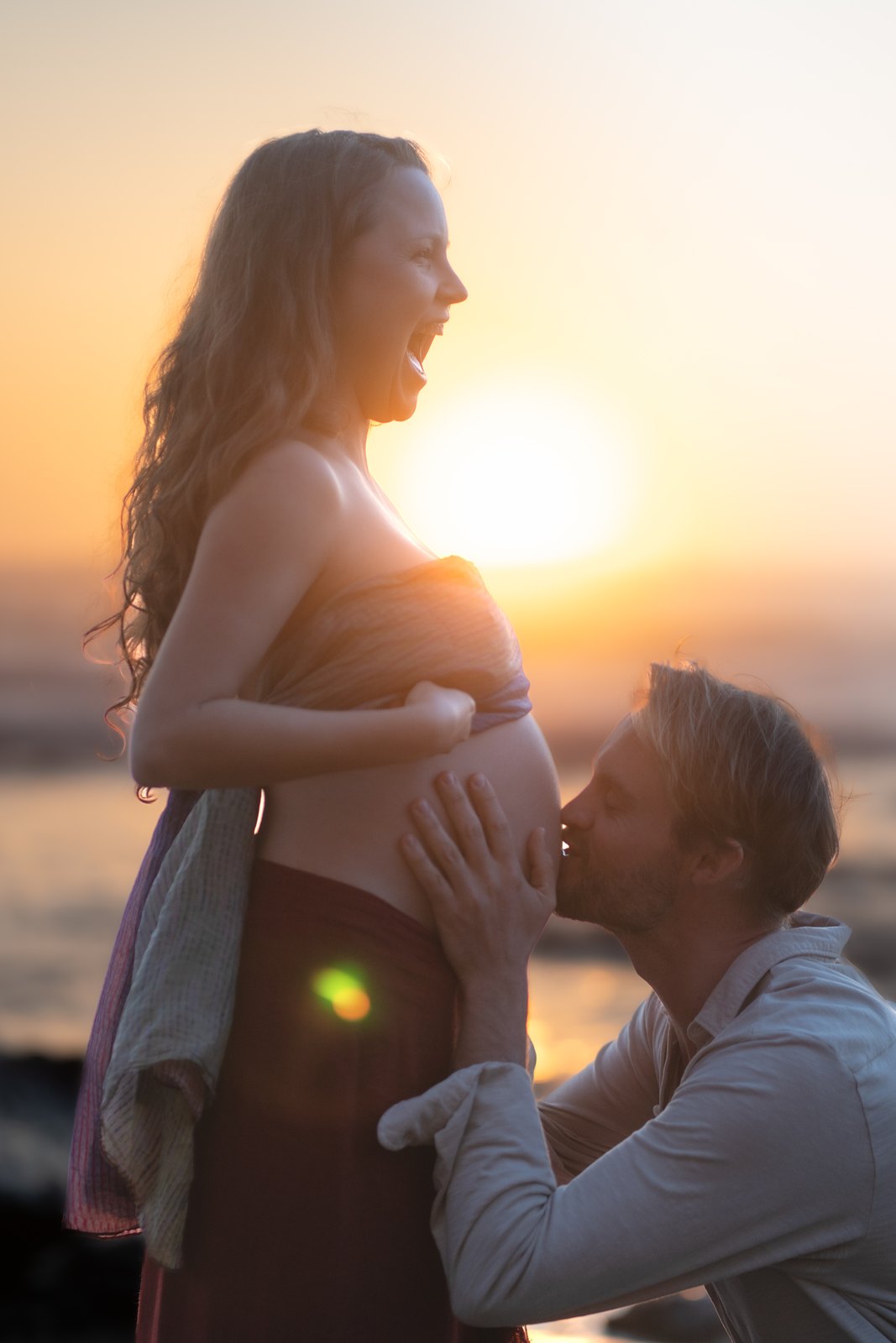 Partner kissing a pregnant belly at sunset